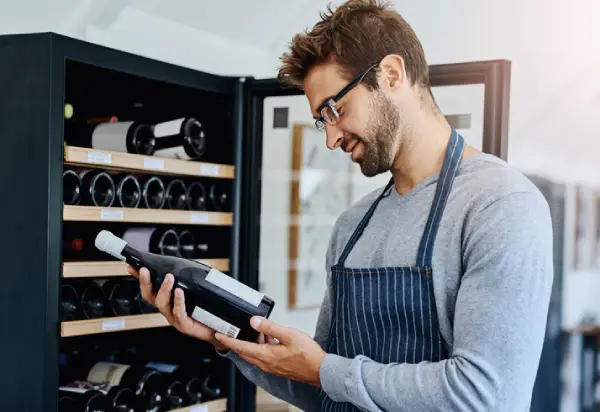 Man wearing a blue apron and holding a bottle of wine while standing in front of a single-zone wine cooler.